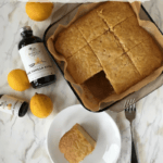 easy lemon cake with a corner piece served on a small plate, with some lemons and bottles of Rodelle extracts