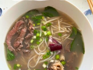 close up of a large bowl of Vietnamese style beef pho with rare beef, bean sprouts, red onions, scallions, rice noodles, and cooked beef.