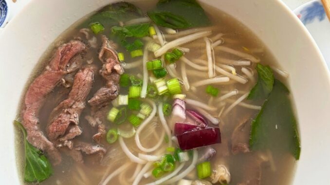 close up of a large bowl of Vietnamese style beef pho with rare beef, bean sprouts, red onions, scallions, rice noodles, and cooked beef.