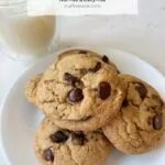 a pile of olive oil chocolate chip cookies on a white plate, next to a glass of milk