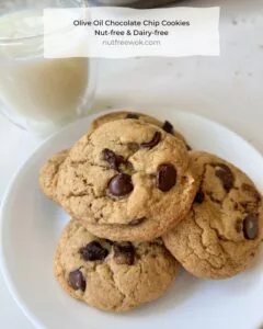 a pile of olive oil chocolate chip cookies on a white plate, next to a glass of milk